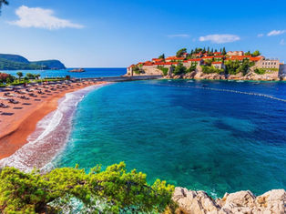 Wide view of Sveti Stefan, Montenegro, showing the historic island resort, turquoise Adriatic Sea, and a curved sandy beach with rows of sun loungers.