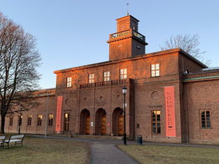 Brick building with trees and benches in foreground. Two red banners read "VIGELAND JUBILEET." Clear sky, warm sunset glow.