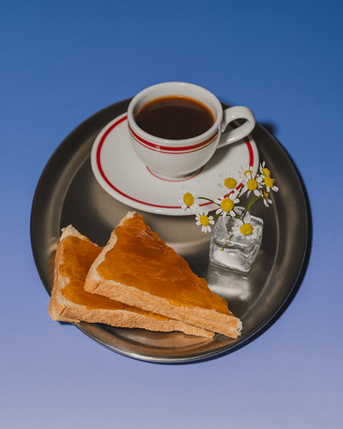 A silver tray with an espresso shot, small vase of flowers and bread spread with jam are pplaced in a studio setting with blue background.