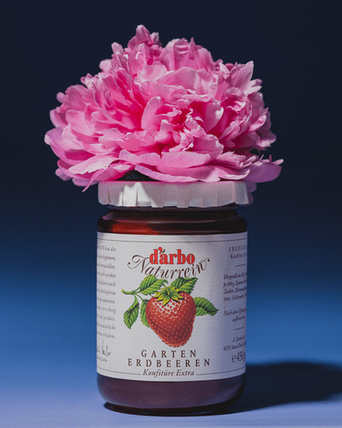 A glass of strawberry jam is standing in the studio, label facing the camera.  A pink peony flower is placed on top of the glass.