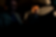 Two people in a dimly lit car, one driving intently, the other in shadow. Tense mood with a dark, rainy background.
