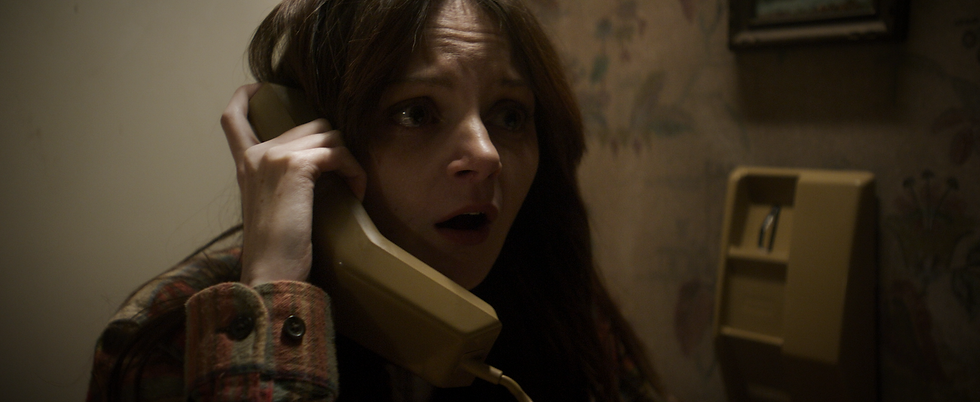 A worried woman with open mouth holds a beige rotary phone. She's in a dimly-lit room with floral wallpaper, wearing a plaid jacket.