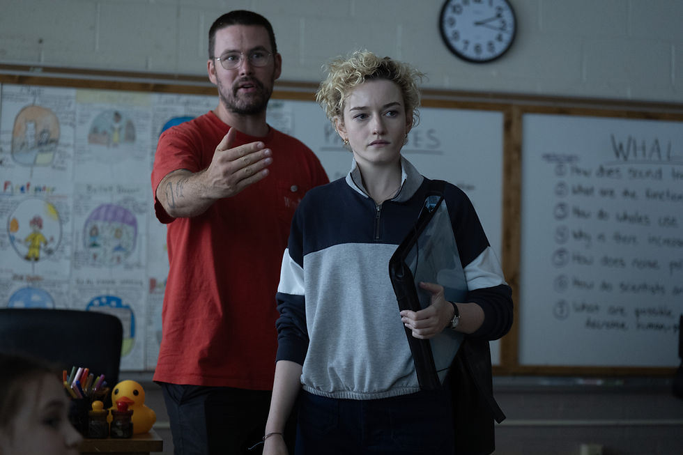 Two people in a classroom, one gesturing. A board with notes and drawings in the background. A clock on the wall shows the time.