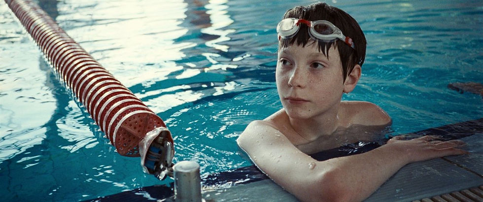 Young boy in swim goggles rests at edge of indoor pool, near a red lane divider. Water reflects blue, creating a calm mood.