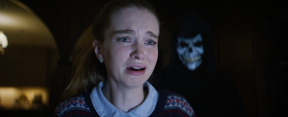 Woman in fear wearing a festive sweater with a skull-masked figure behind her in a dimly lit room, creating a tense atmosphere.