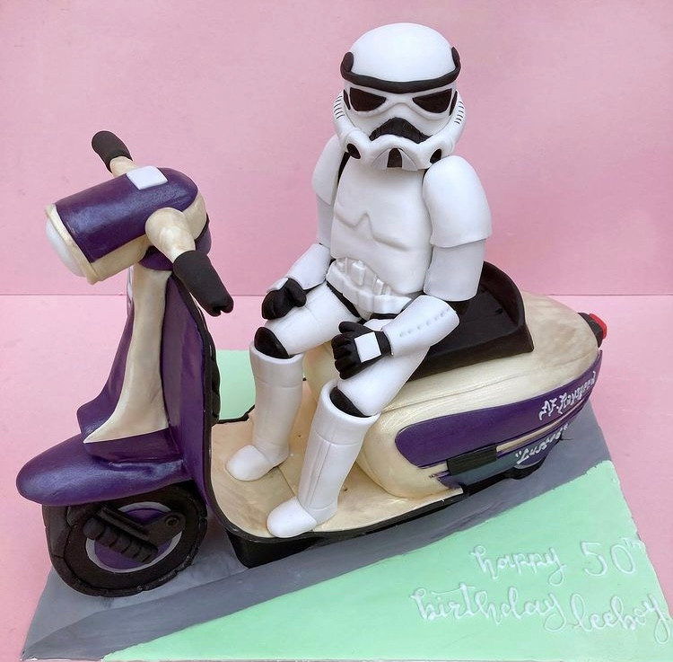 Image shows a cake carved into the shape of a purple moped with a Star Wars Stormtrooper, made from fondant, sitting on it.