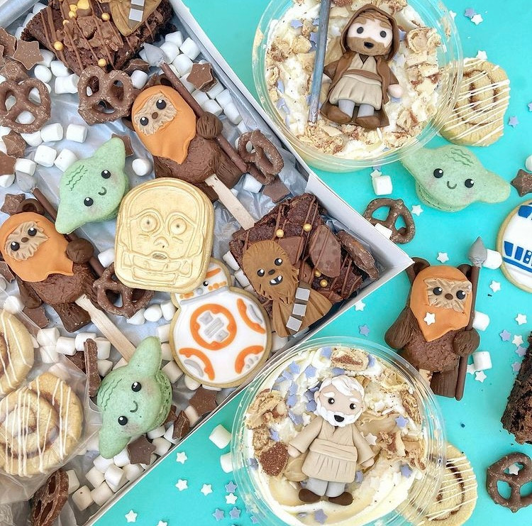 Image shows the contents of a Star Wars treat box. It includes cake with Hans Solo and Obi Wan, Yoda macarons, Chewbacca brownies and other Star Wars related treats.