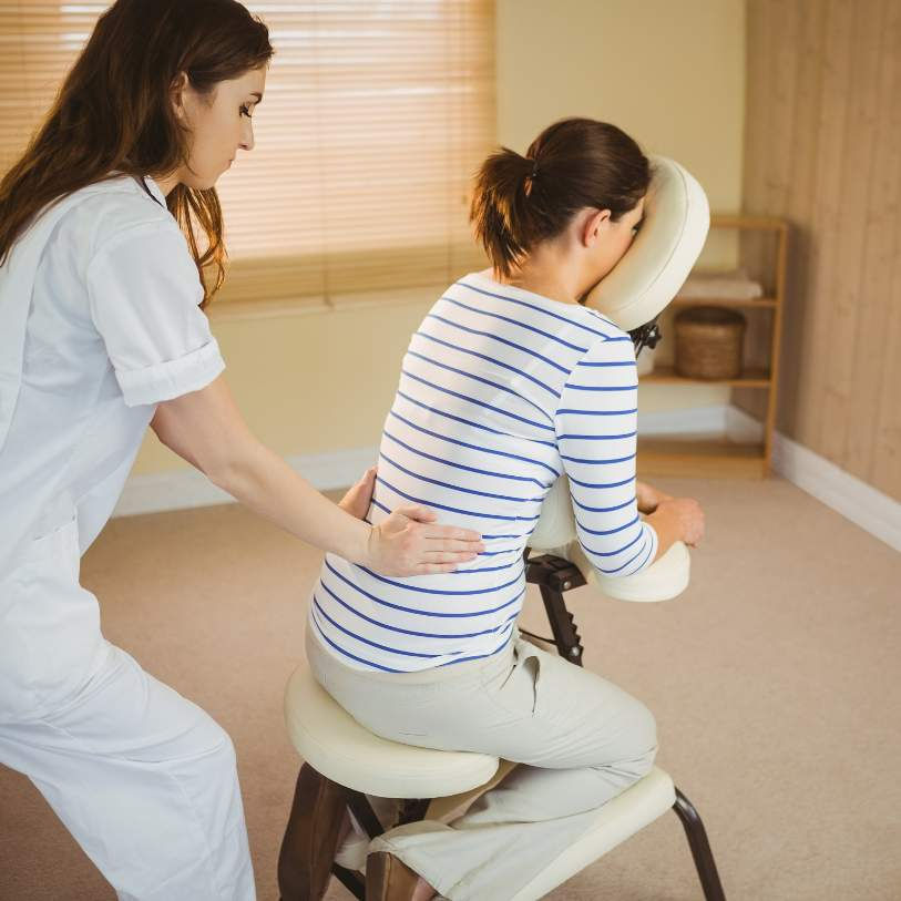 "Female massage therapist providing a relaxing seated back massage to a client in a cozy, well-lit room with wooden decor."