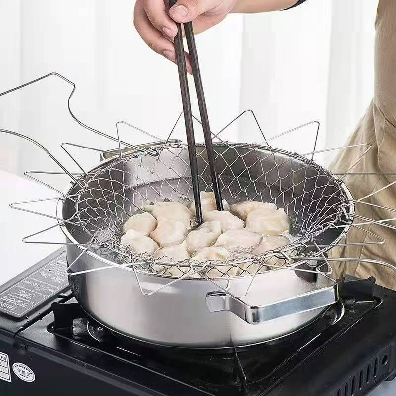 cesto de cozinha dobrável em metal, sendo usado para cozinhar ou fritar alimentos (parecem ser bolinhos ou pastéis) dentro de uma panela no fogão. Uma pessoa usa hashi para mexer os alimentos dentro do cesto.