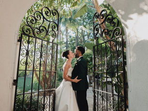 A bride and groom embraces romantically under a wrought iron archway, framed by lush greenery.