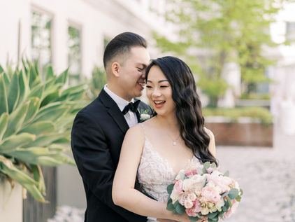 A couple embraces joyfully outdoors. The bride holds a pink bouquet, wearing a lace gown; the groom in a black suit. Greenery surrounds them.