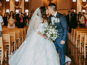Bride and groom kiss in a church aisle. The bride wears a white gown and veil, holding flowers. Guests watch from wooden pews in the background.