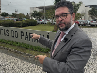 Autor de terno apontando para a placa de entrada do tribunal de contas do estado do paraná