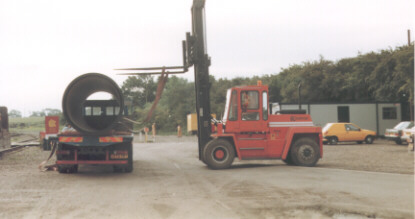 Forklift truck stands alongside lorry flatbed loaded with large pipes in railway yard