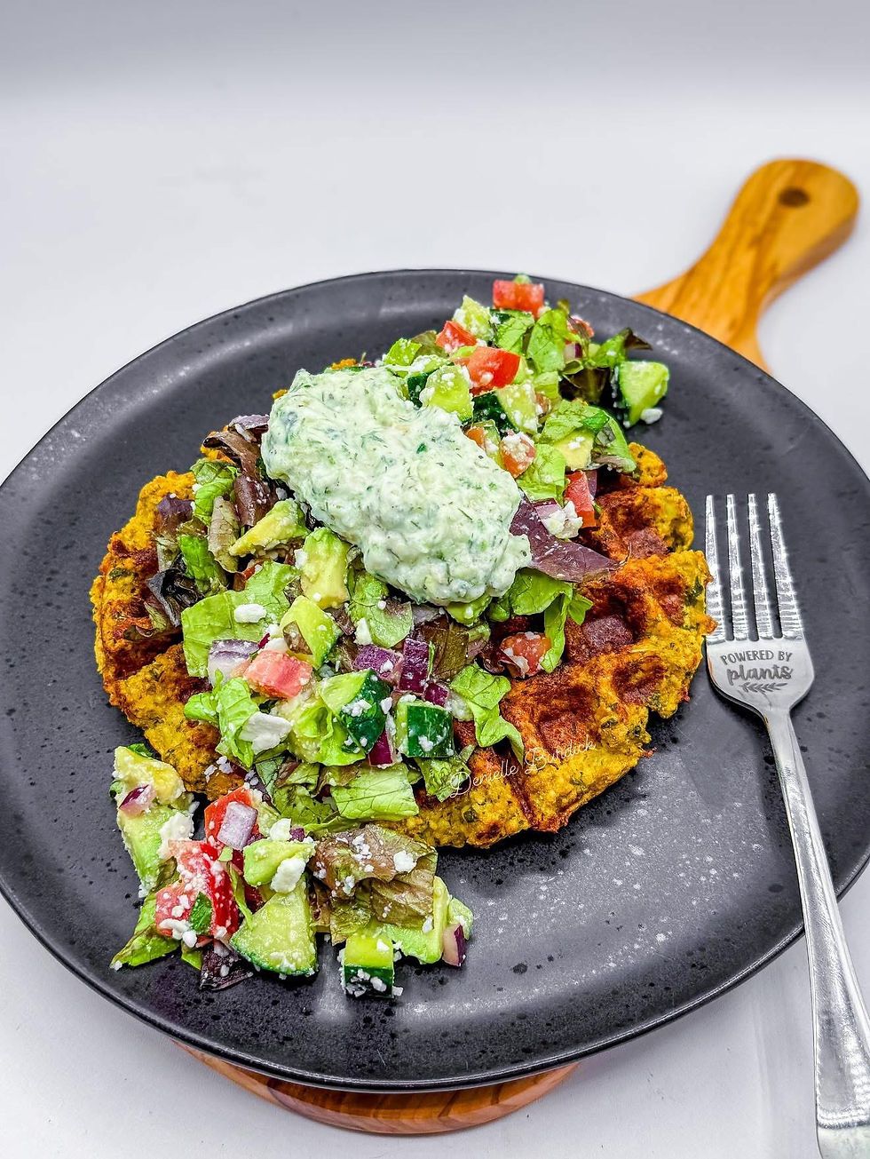 Waffle topped with vibrant salad and dollop of green sauce on a black plate. Fork with "Powered by Plants" text on wooden board.