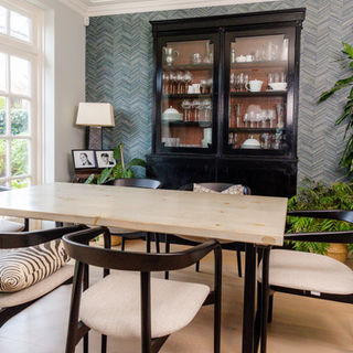Dining area with black dining chairs washed grey wooden table.  There is an antique black library cabinet stacked with glasses and white china.  On the walls is zig zag textured wallpaper in teal and large plants in each corner.  Traditional white patio doors and oak floor.
