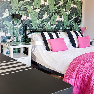A bedroom with banana leaf wallpaper behind the bed, a black and white stiped rug, chest of drawers and cushions on the bed. There is a bright pink throw and cushions on the bed.