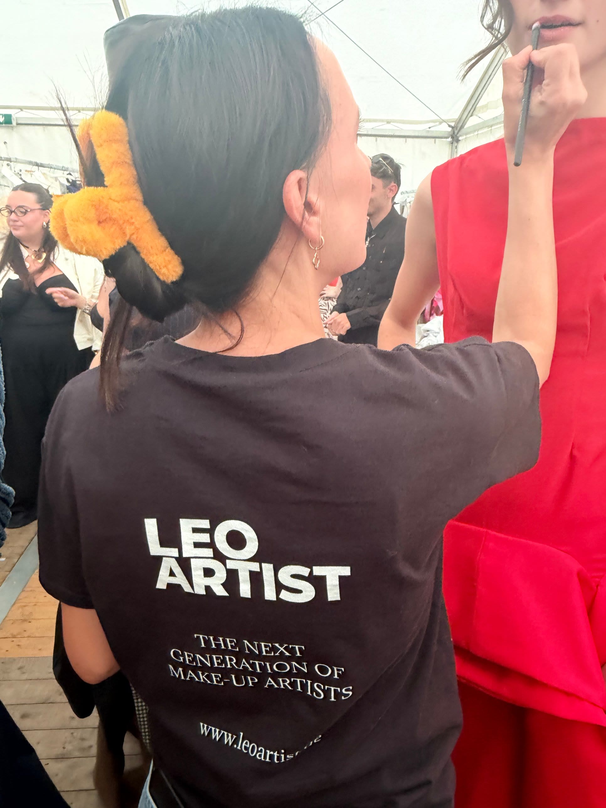 Schülerin im Leo Artist Shirt schminkt backstage ein Model im roten Kleid
