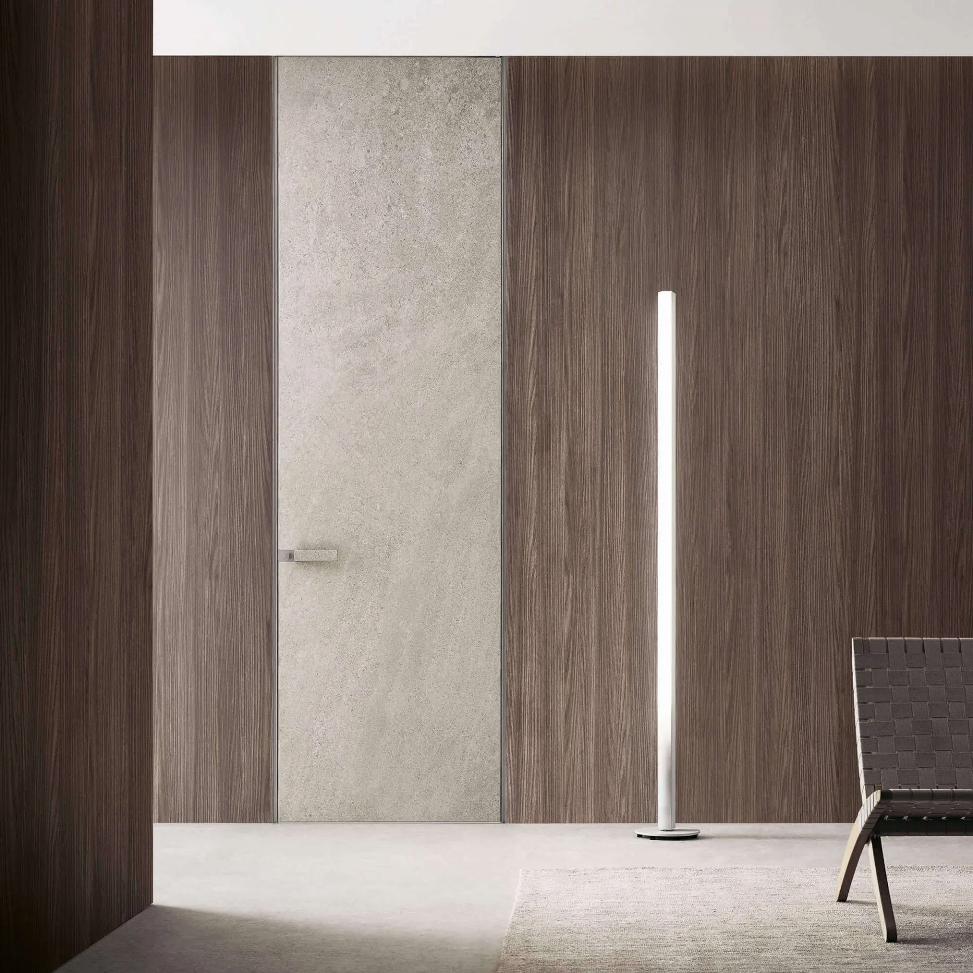The second photo shows the same door in a broader context. The door is flush with the wooden wall, and its concrete finish blends in perfectly with the modern décor of the room. The interior is decorated in neutral, earthy tones, with modern accents, such as a slim, minimalist floor lamp.