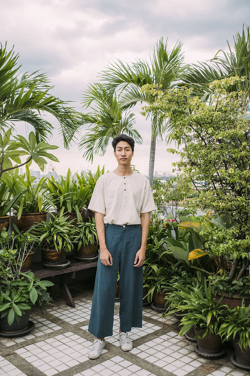 A guy standing against a green back drop of plants wearing green trousers and a white t-shirt tucked in