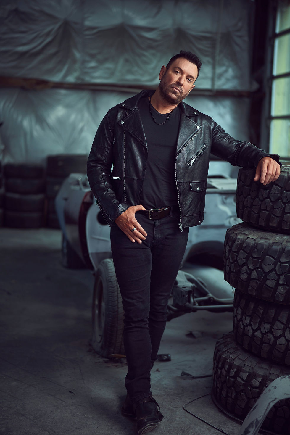 Scott Pryor in a black leather jacket leans on stacked tires in a garage. Background shows car parts and a draped wall, creating a rugged vibe.