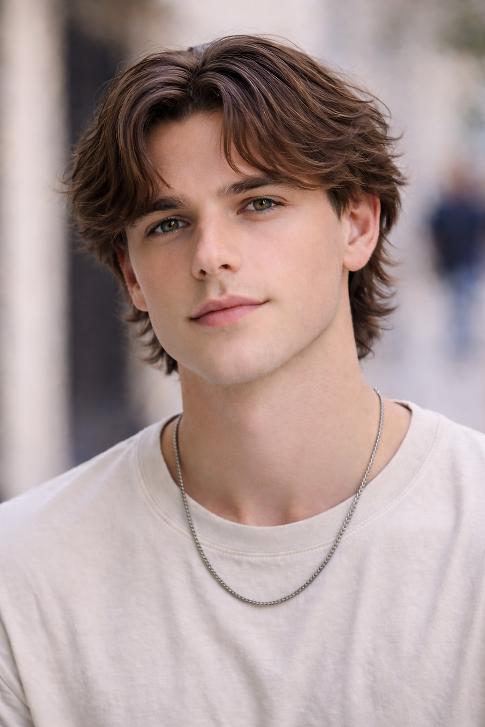 Young man with brown hair and a neutral expression in a white shirt and necklace. Blurred urban background adds a relaxed mood.