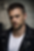 Man in a black leather jacket and white shirt, looking serious against a blurred gray brick wall background.