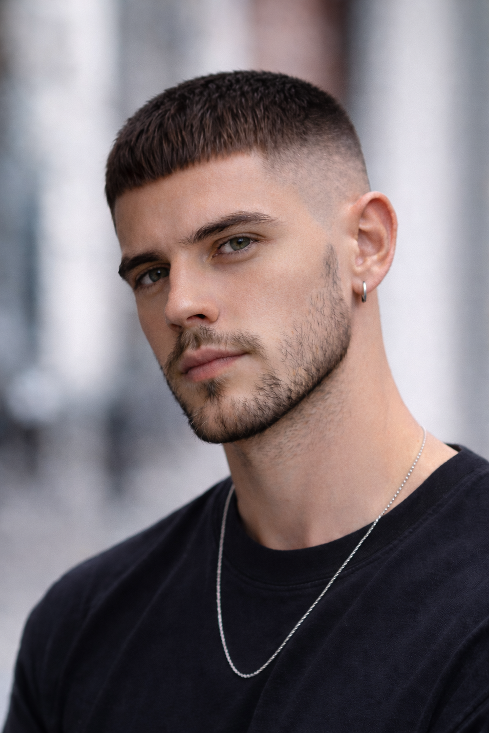 A man with short hair and an earring gazes intently. He wears a black shirt and silver chain. Blurred background suggests an urban setting.
