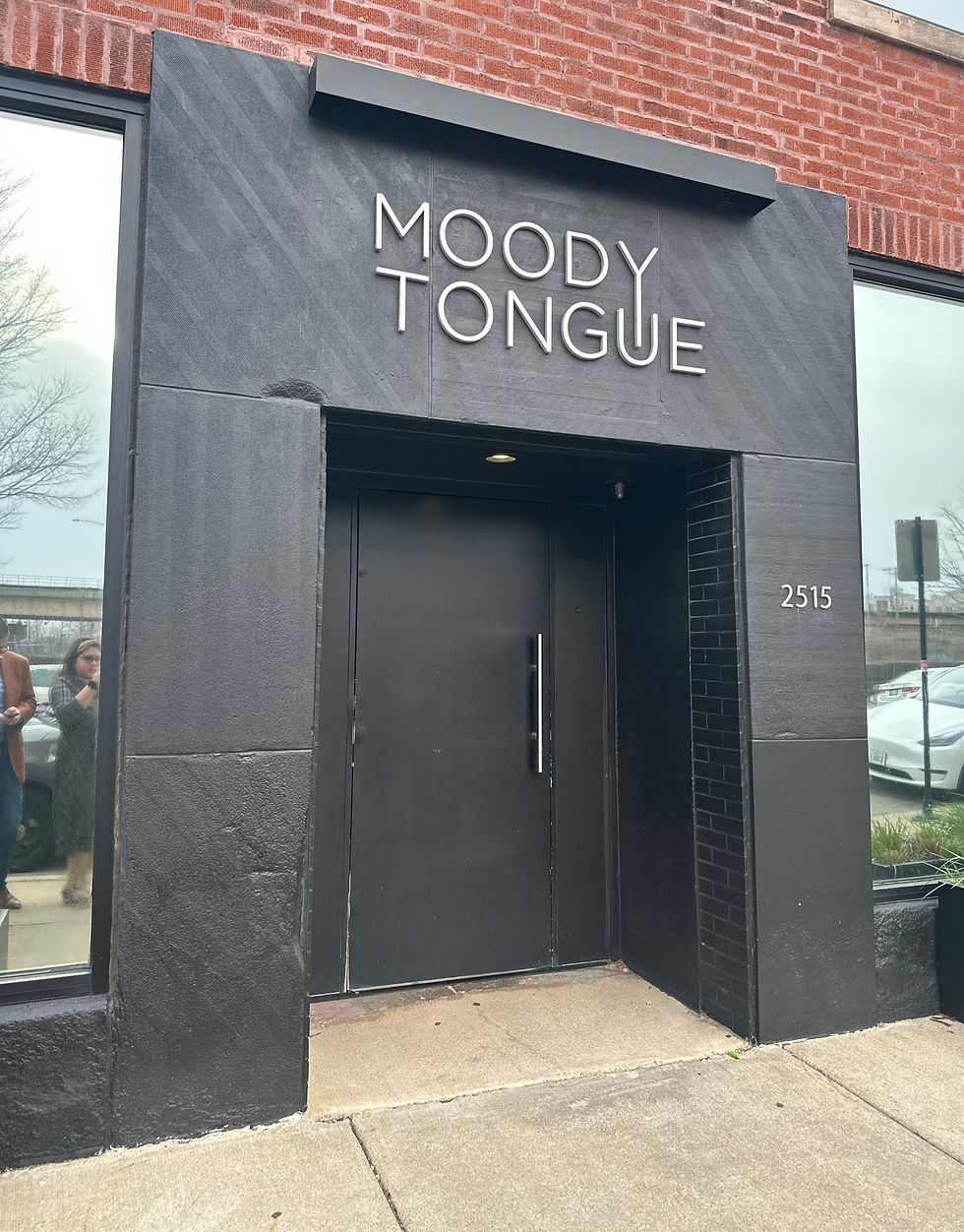 Black entrance with "MOODY TONGUE" sign, surrounded by red brick. Reflections of people in window. Address "2515" visible. Cloudy day.