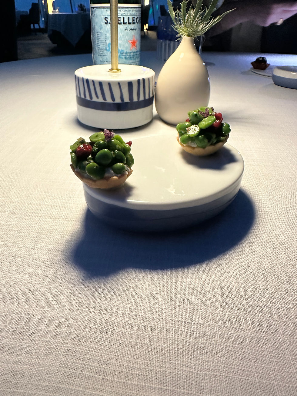 Two small pastries with green and red toppings on a white plate. A vase with plant and a S.Pellegrino bottle in a dimly lit setting.