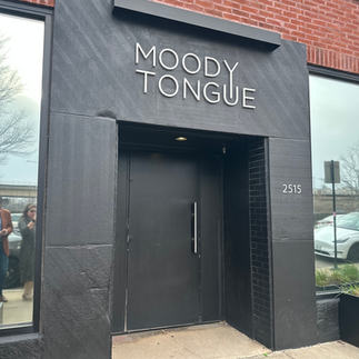 Black entrance with "MOODY TONGUE" sign, surrounded by red brick. Reflections of people in window. Address "2515" visible. Cloudy day.