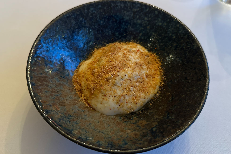 A textured dark bowl holds a Rabbit Confit dish topped with brown spices, set on a white tablecloth. The bowl's interior has a blue hue.