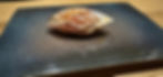 Grilled sushi on a dark ceramic plate, showcasing pink fish with charred lines, set on a wooden table under soft lighting.