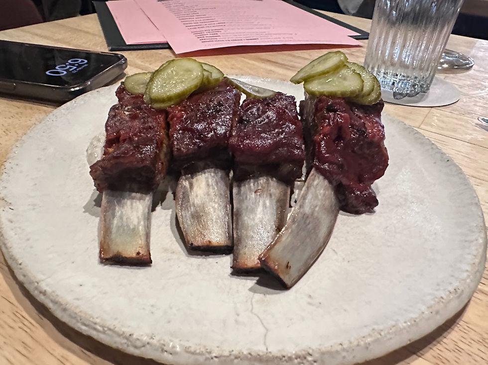 Beef ribs topped with pickles on a white plate, set on a wooden table. A smartphone shows 9:54, with menus and a water glass nearby.