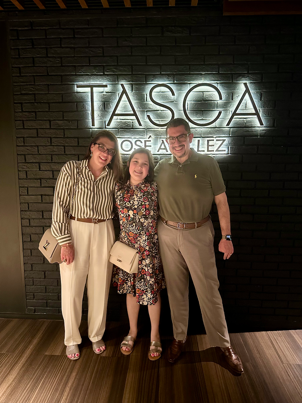 Three people smiling, posing in front of a wall with "TASCA" text. They're wearing stylish outfits in a warmly lit setting.