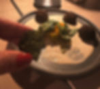 Plate of falafel with tahini, garnished with herbs and vegetables. A hand with red nails holds a piece. Warm, dimly lit setting.