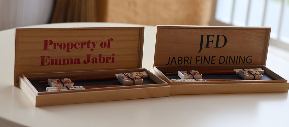 Two wooden boxes on a table, each holding a pair of decorated chopsticks. Texts read "Property of Emma Jabri" and "JFD Jabri Fine Dining."