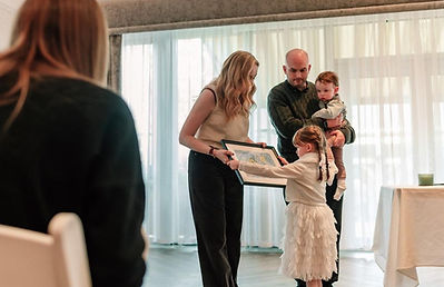 A mother is passing a framed drawing to young girl. The girl drew the picture for her brother. It is her brother's naming day. Beside them the father holds his son, whose naming day it is.
