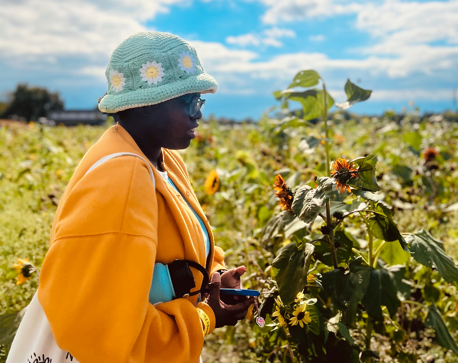 Nosa in a yellow cardigan and a flower patterned bucket hat. She has a water bottle under her arm and a tote bag hanging from her shoulder. In her hand is her phone with a charm hanging from it. She is looking to the right and facing a tall sunflower plant.