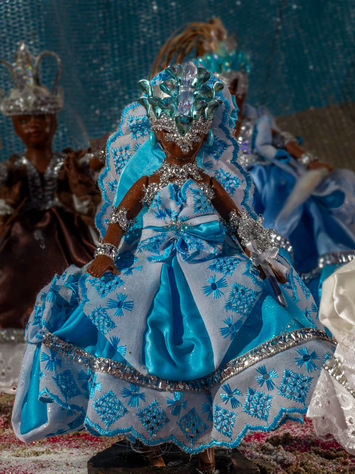Uma boneca de plástico de pele negra está em pé sobre um pedestal. Ela usa um vestido azul claro, todo bordado com laise branca. Seu rosto está encoberto por uma espécie de máscara que envolve também os seus cabelos. Outras bonecas, que representam outros orixás, podem ser vistas parcialmente ao seu lado e atrás dela.