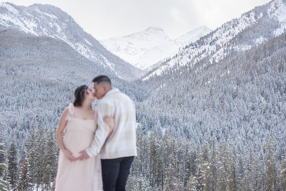 A couple embraces and kisses in front of a snow-covered mountain backdrop. She wears a light dress, creating a serene and romantic scene. Invermere Maternity Photographer