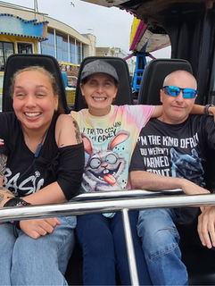 A man and two women in a theme park cart with their arms around each other. All three are smiling at the camera.