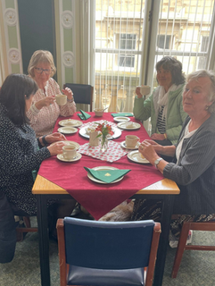 A group of Volunteers sat around a table in the Town Hall having scones and tea. They are all looking at the camera and smiling.