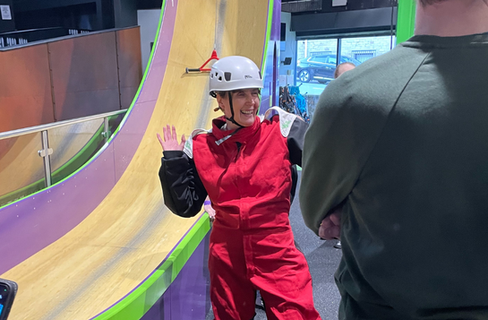 A lady in a Red jumpsuit with a White helmet on. The lady is stood on crash mats and appears to be attached to a cord.