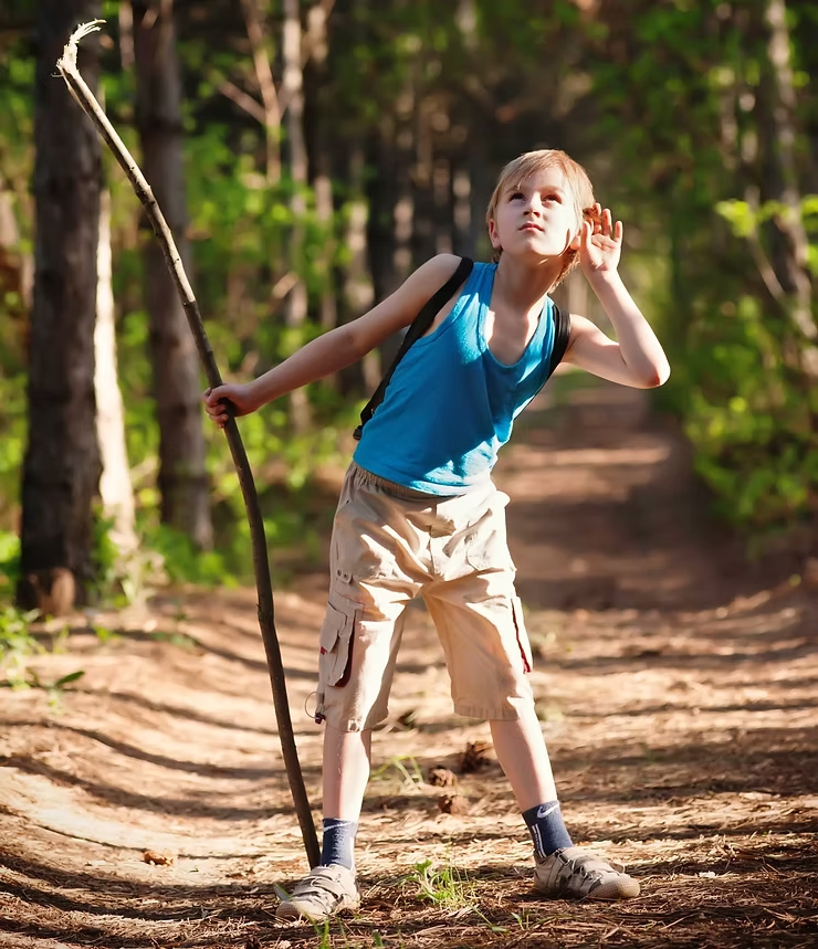 Child in blue tank top holds a stick, listening intently in a sunlit forest path. Green foliage, relaxed mood, natural setting.