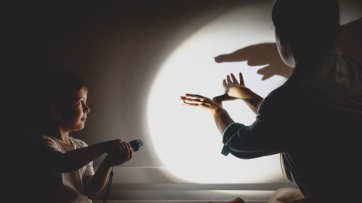 Child holds flashlight, casting shadow puppet on wall with adult. Dimly lit room, focused expressions.