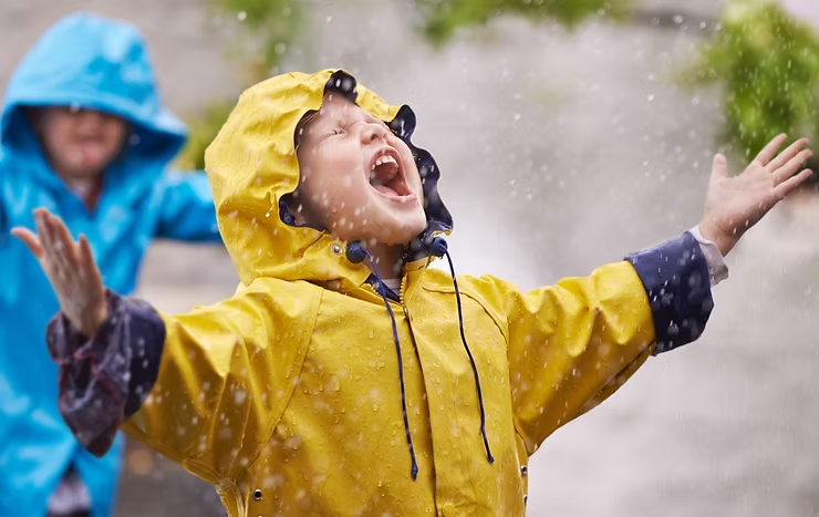 Children in yellow and blue raincoats joyfully playing in the rain, arms outstretched, with water droplets visible around them.
