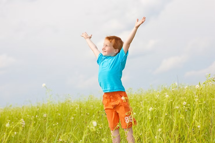 Child in blue shirt and orange shorts stands in a green field, arms raised joyfully, under a cloudy sky, expressing happiness.