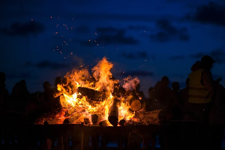 A bonfire with bright flames against a dark blue evening sky. Silhouetted people are gathered around.