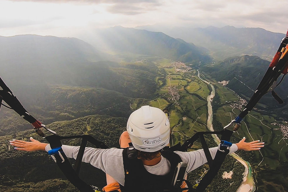 Person paragliding over green Soča Valley, arms outstretched. Wearing a white helmet. Mountains and river visible. Dramatic clouds above. Ton of Fun Paragliding. Soča Valley, Slovenia.